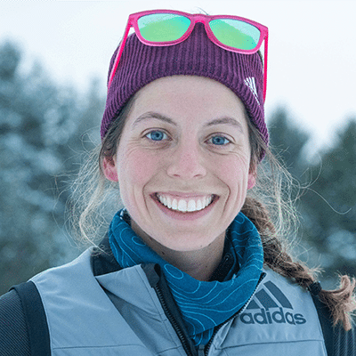 corrine smiling wearing pink sunglasses on top her head, a purple stocking hat and a gray adidas coat