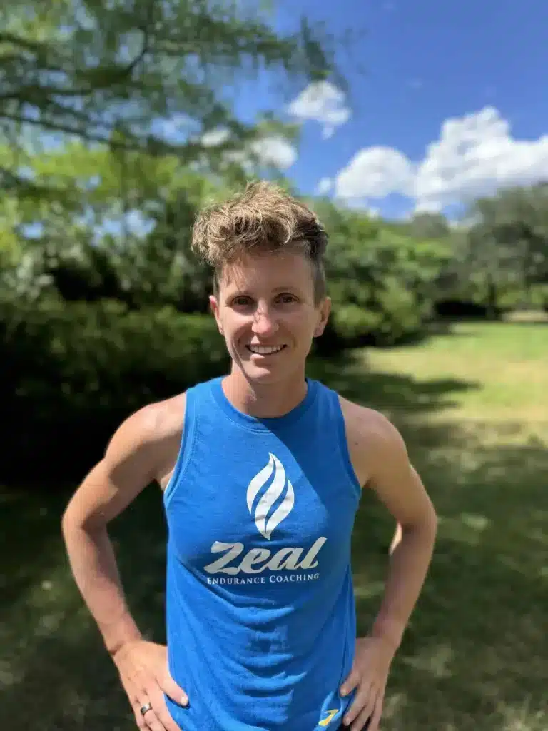 Dee smiling with trees and a blue sky, wearing a blue Zeal Endurance Coaching tank top with hands on hips