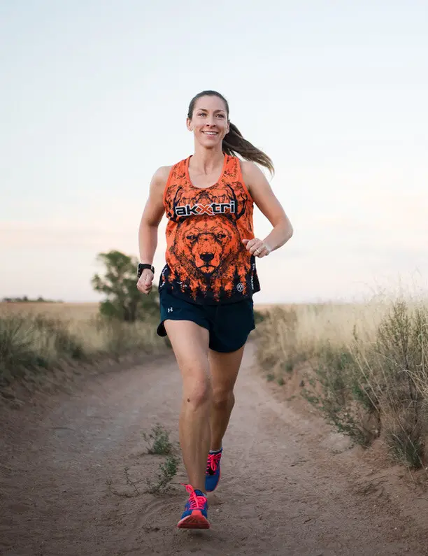 kelly running down. a dirt path in colorful blue and onk shoes and is wearing an orange tank top with akxtri and a bear