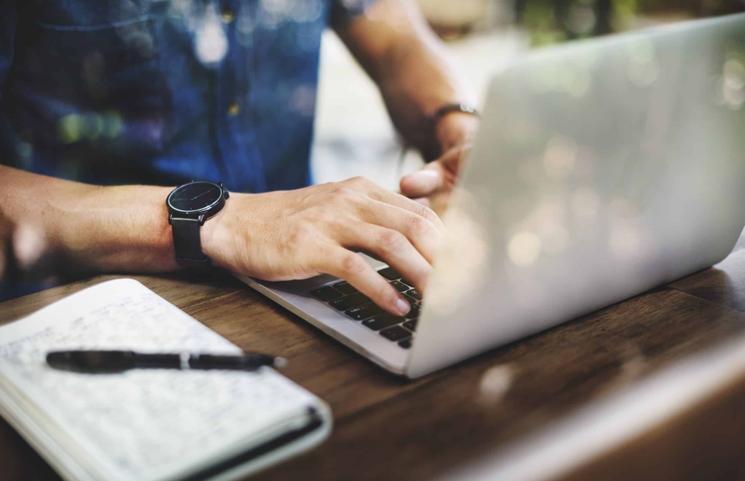 A person typing on a laptop with a notebook and pen next to it