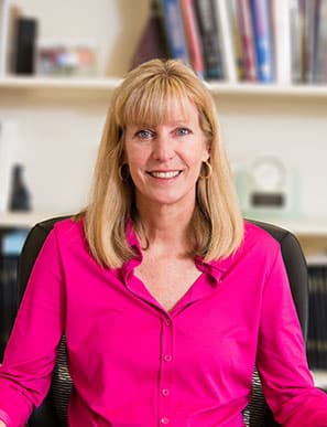 shala smiling with books in the background. she is wearing a pink button shirt and has blonde hair and blue eyes