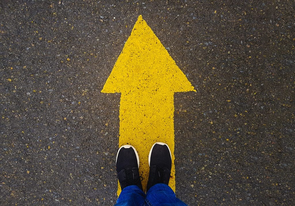 A yellow arrow painted on the asphalt street with two feet standing on top of it