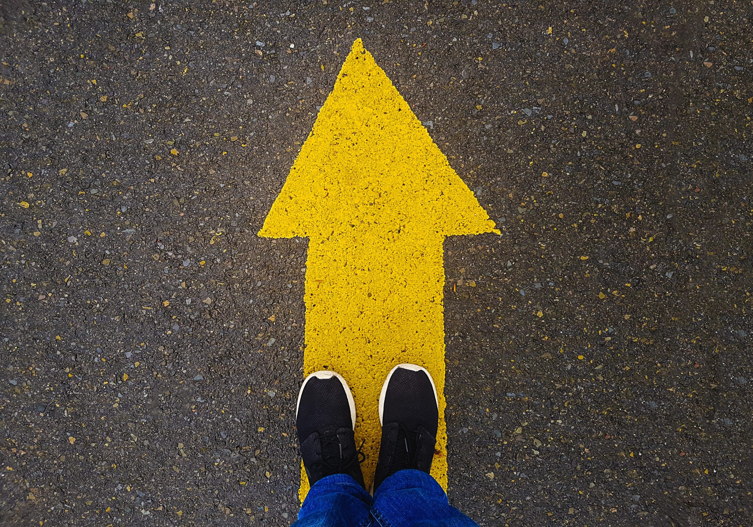 A yellow arrow painted on the asphalt street with two feet standing on top of it