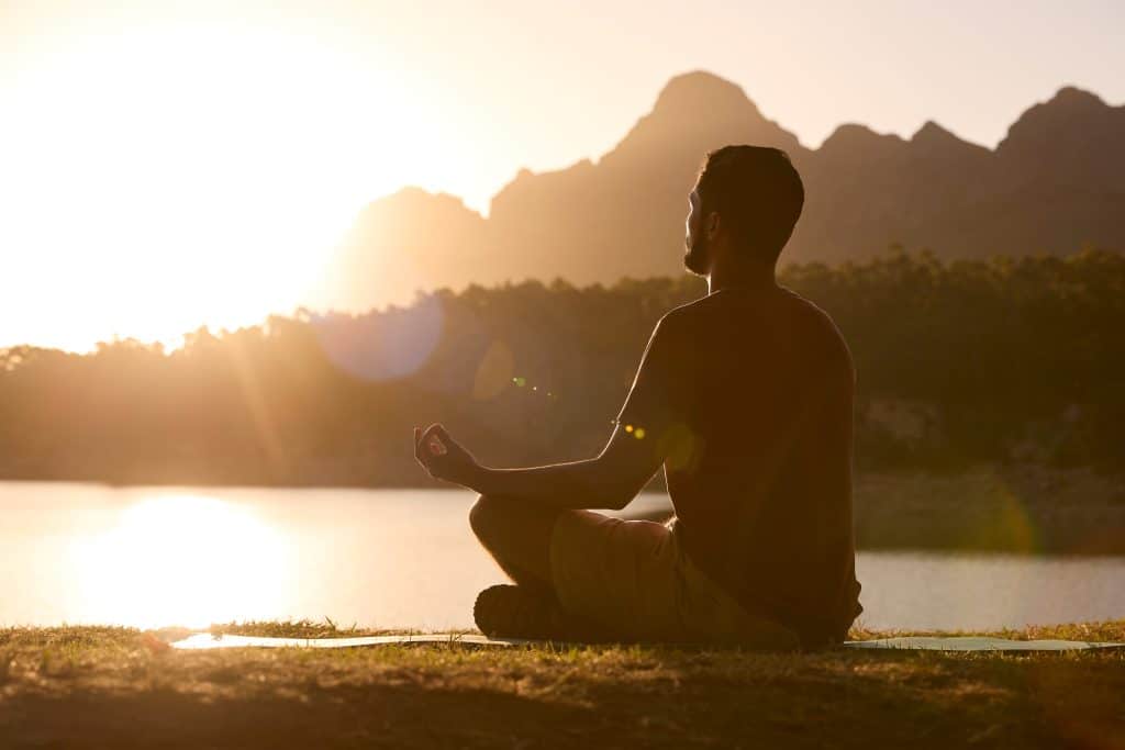 The profile of a man meditating sitting outside next to a lake with mountains in the background