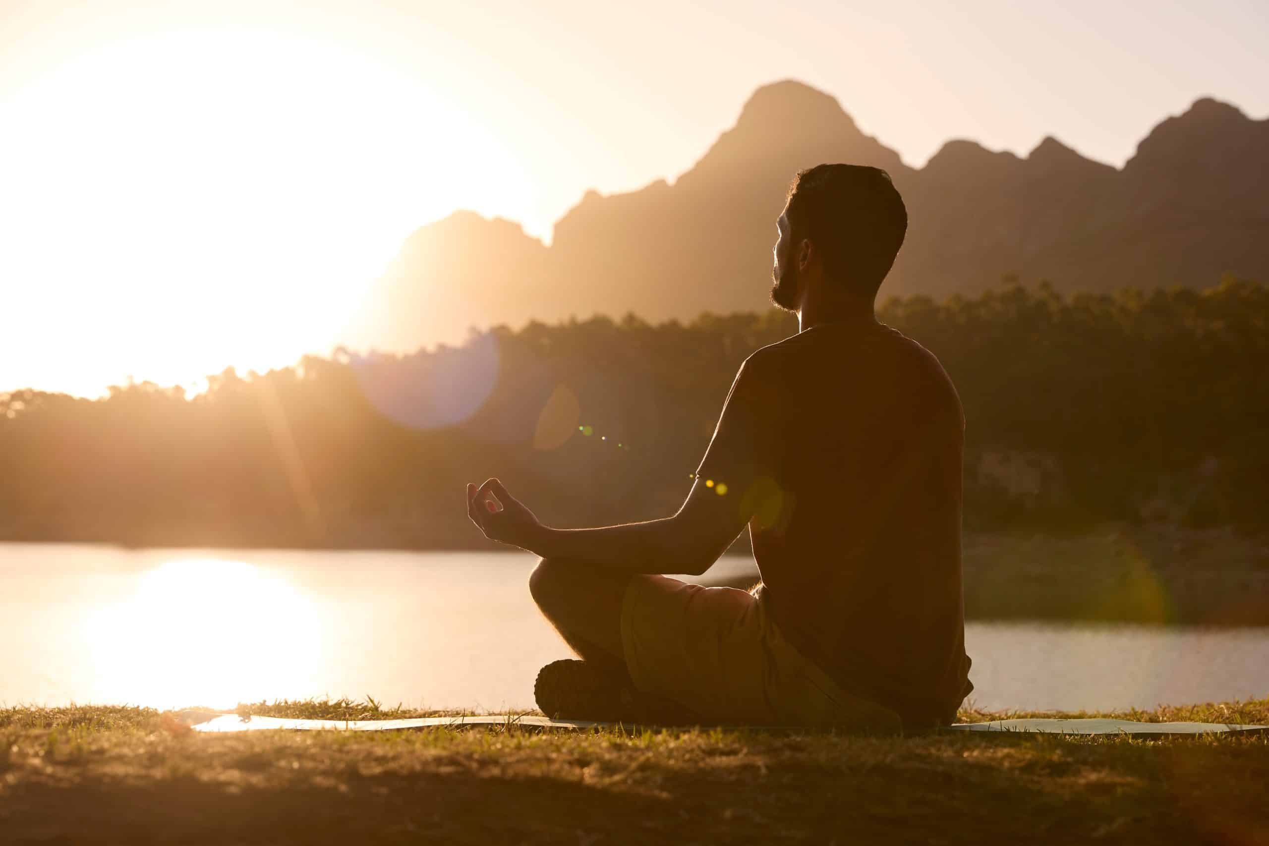 The profile of a man meditating sitting outside next to a lake with mountains in the background