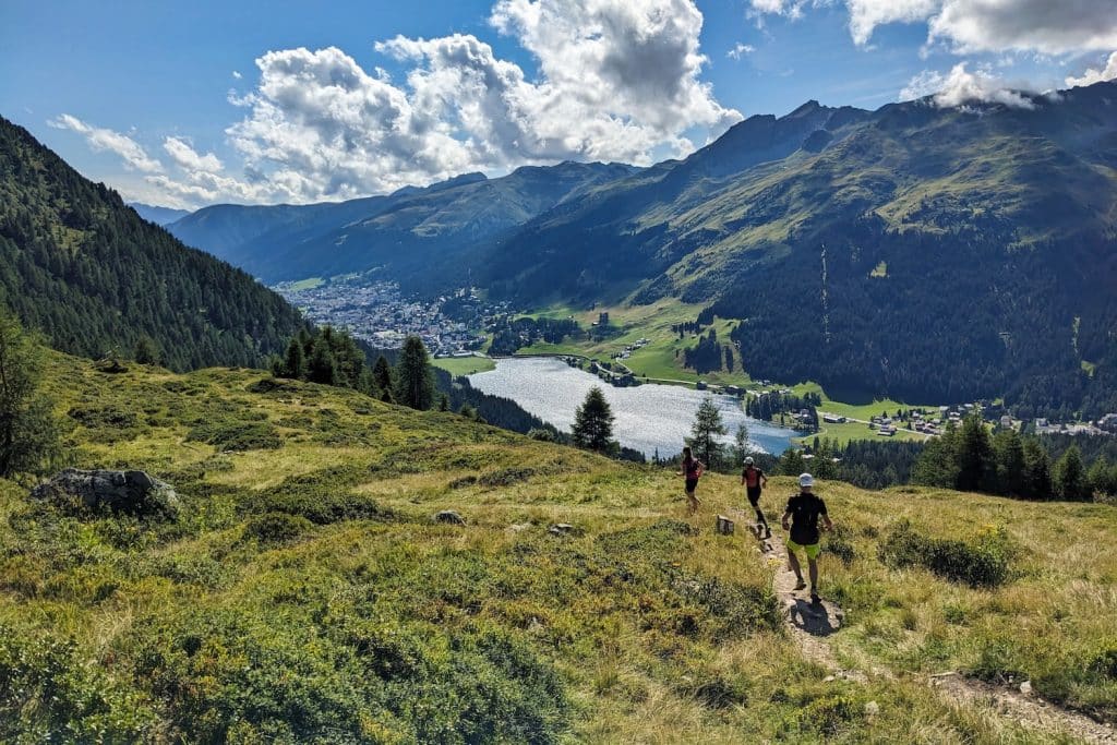 Several trail runners running along a narrow trail down a mountain with green grass on either side and a small lake and a mountain view in the distance