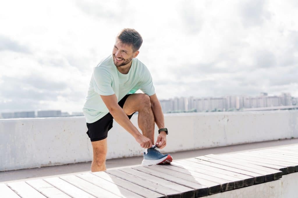 A man in a T-shirt, running sports shoes and smart watch, tying his shoe on a bench outside