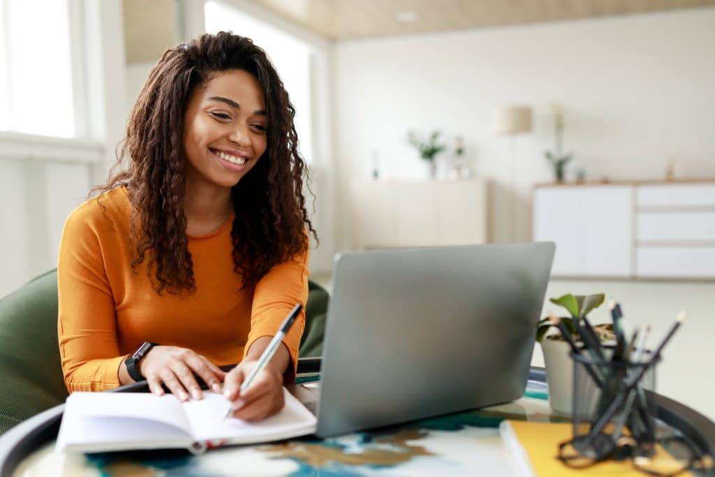 Smiling young African American woman sitting at desk working on laptop taking notes in notebook, happy millennial female studying online, watching webinar using computer and writing check list
