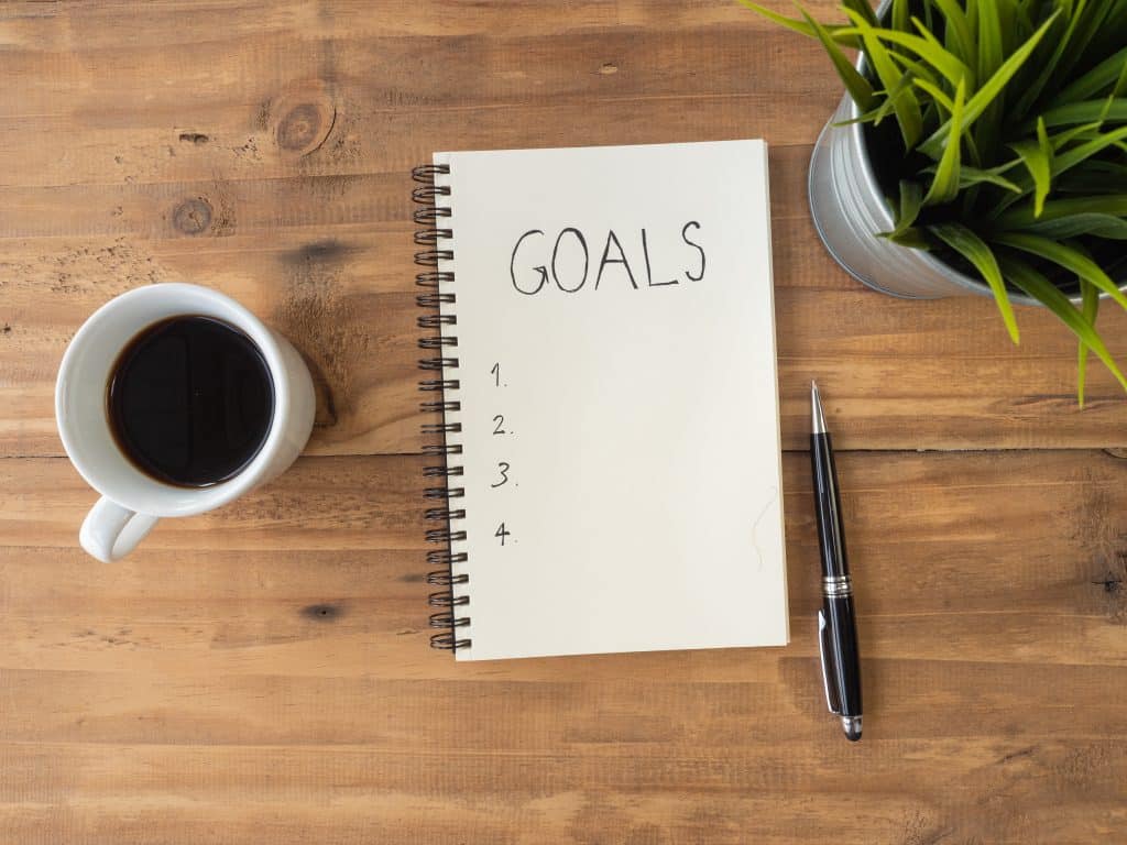 A pad of paper with "goals" written on it next to a cup of coffee on a wooden table