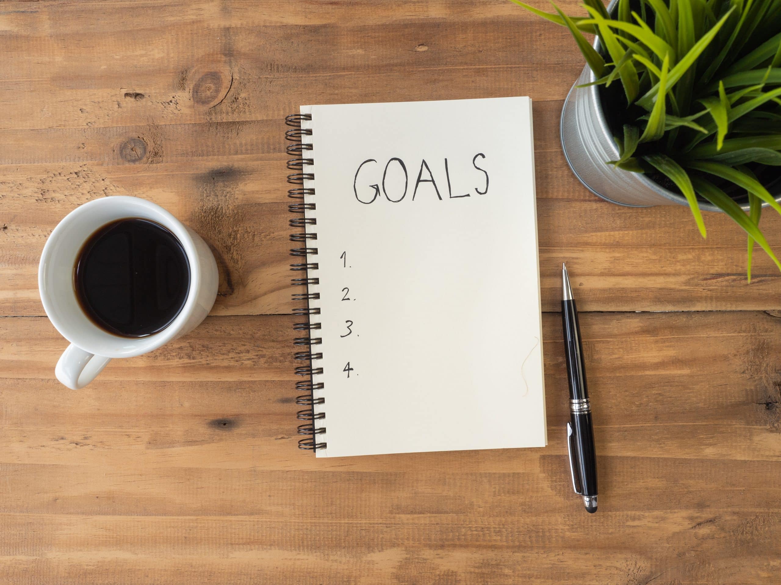 A pad of paper with "goals" written on it next to a cup of coffee on a wooden table