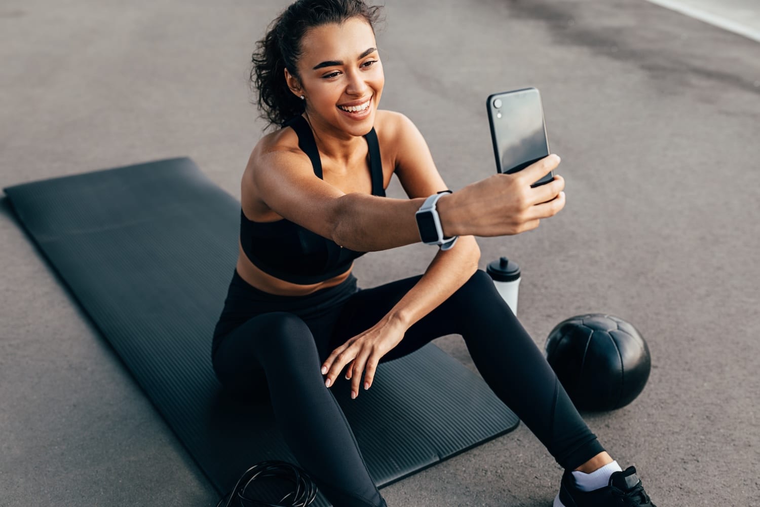 A woman in workout apparel sitting on a yoga mat and taking a selfie