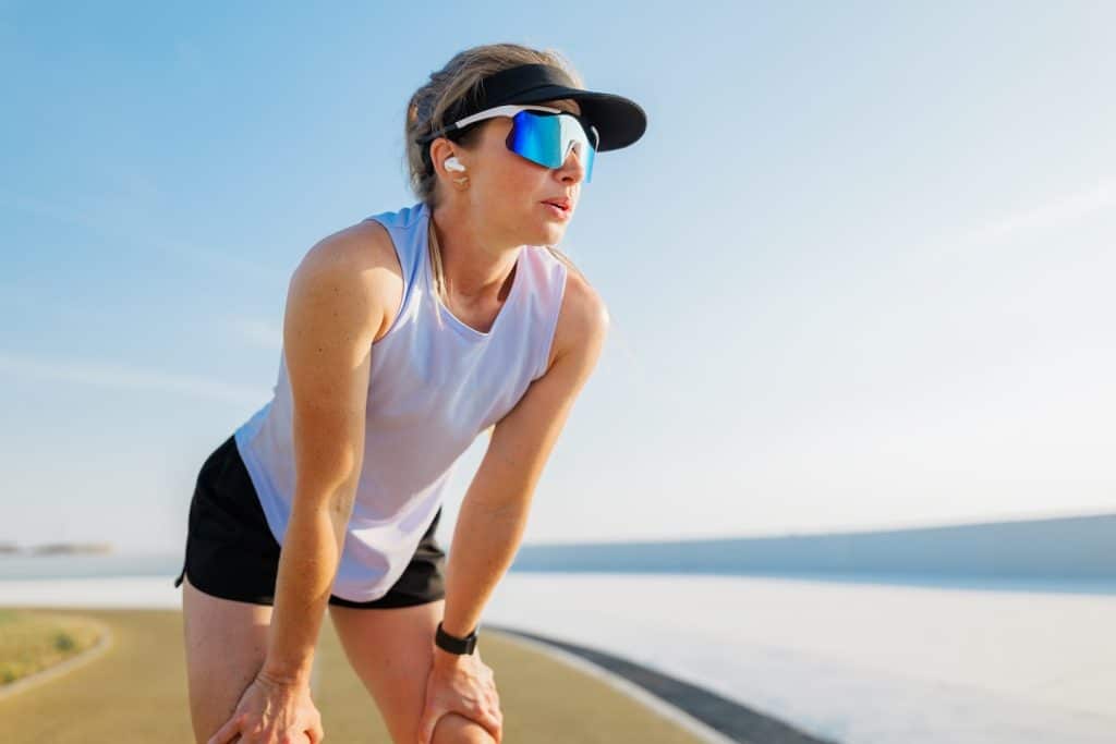 Woman wearing a visor and sunglasses takes a break with her hands on her knees while jogging along a pathway near a body of water on a sunny day