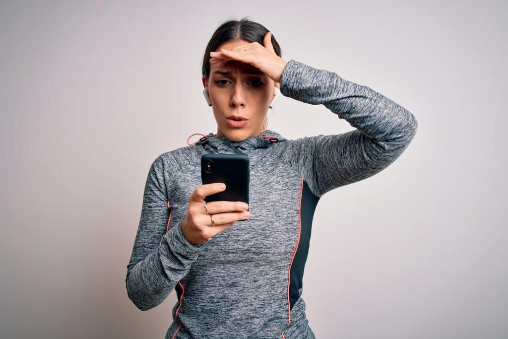 A woman wearing athletic wear using a smartphone and looking stressed with a hand on her head